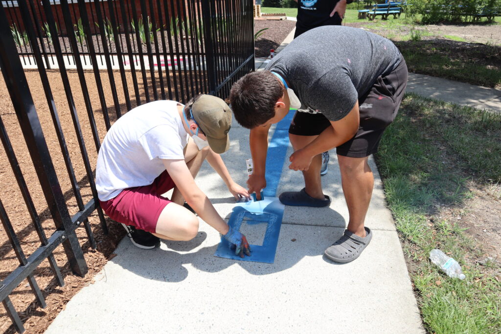 Two volunteers bend over the sensory sidewalk spray painting. One holds a large stencil while the other sprays blue paint.