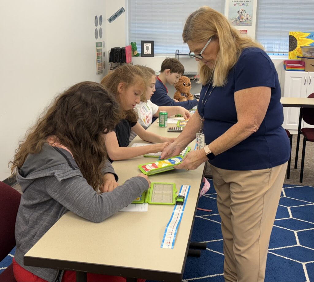 A teacher stands next to a table of students looking at individual measuring tapes and papers.