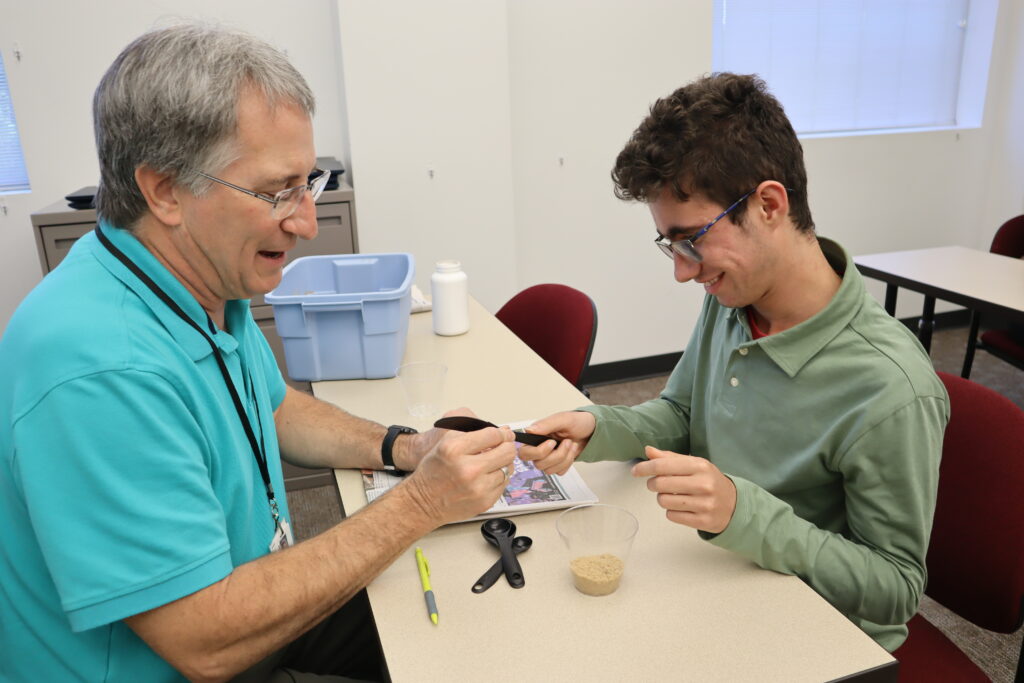 A teacher and student sit at opposite ends of a desk, measuring different volumes of sand in cups.