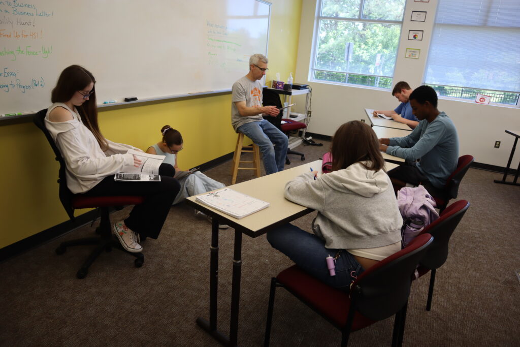 English Class An upper school teacher leads a lesson in front of a classroom with five students reading along to a packet of paper.